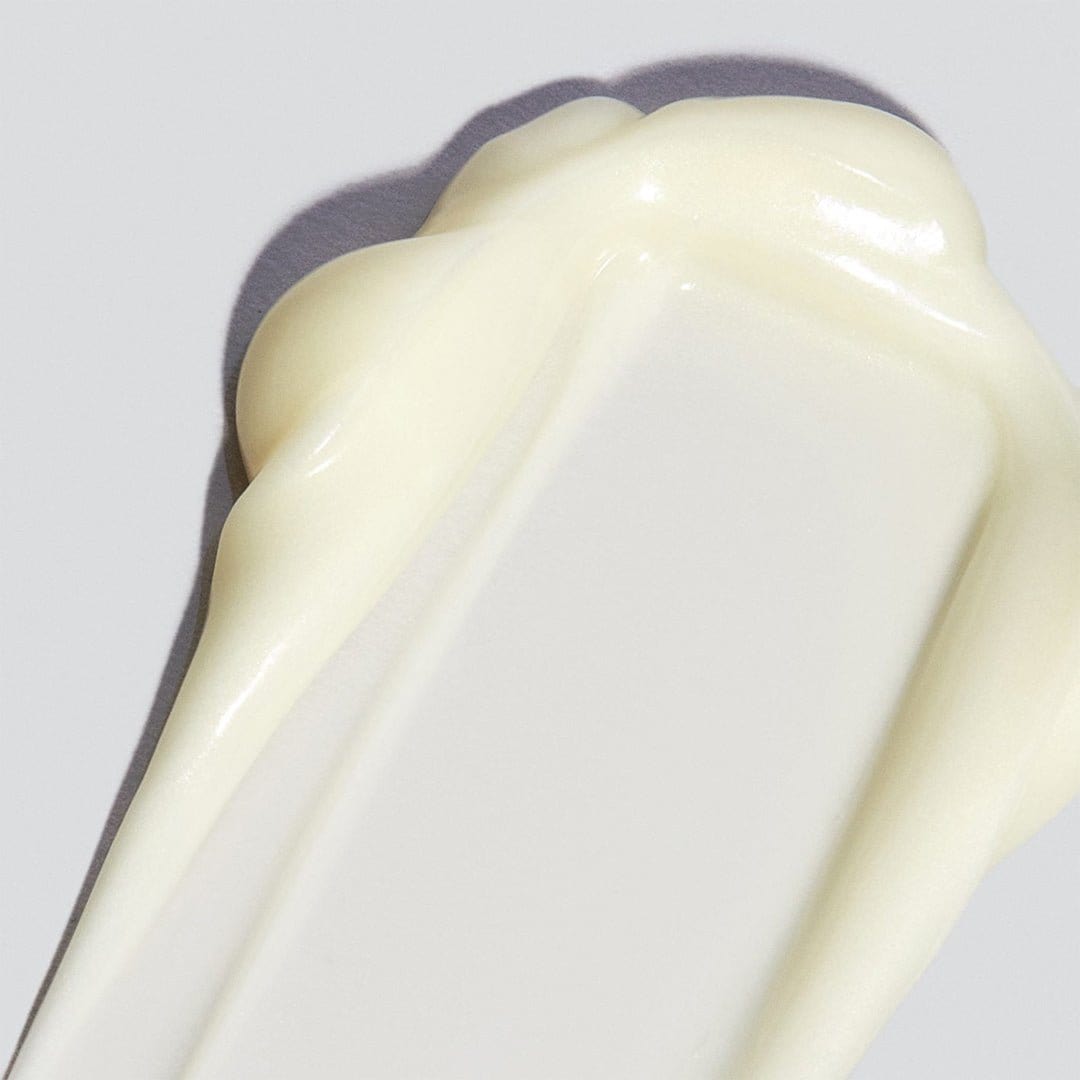 Eine Nahaufnahme eines cremigen, cremefarbenen Abstrichs des AGE Reversal Eye Complex mit Niacinamid, der zur Bekämpfung von Hautalterung entwickelt wurde, auf einer glatten hellgrauen Oberfläche.