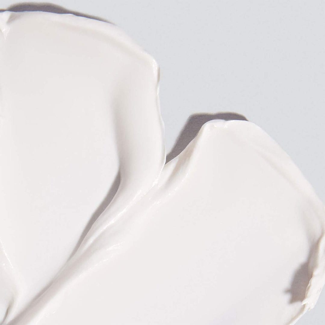 Nahaufnahme von Invisible Physical Defense SPF30, einer glatten weißen Creme mit physischem Sonnenschutz, die in dicken, überlappenden Schichten auf einem hellgrauen Hintergrund aufgetragen ist.