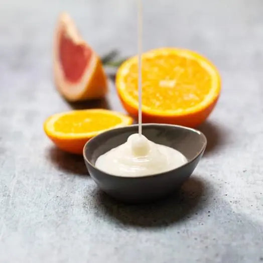 Weiße Pflegetextur fließt in eine graue Schale, dahinter halbierte Orangen und eine Grapefruit