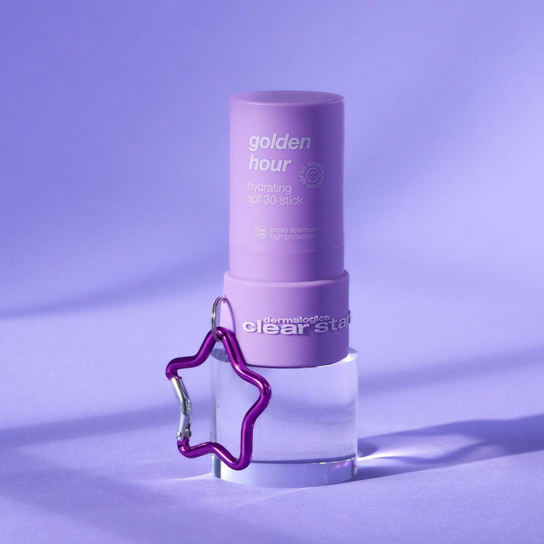 Eine violette Tube mit der Aufschrift "Golden Hour Hydrating SPF 30 Stick" steht auf einem durchsichtigen Sockel, an dem der Clear Start Keychain - gratis befestigt ist, vor einem violetten Hintergrund. Kein Verkaufsprodukt.