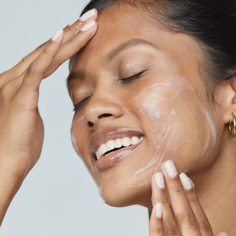 Una persona con los ojos cerrados aplica suavemente la Crema Reparadora Estabilizadora + Limpiador UltraCalmante en su mejilla mientras sonríe: un ritual relajante para pieles sensibles.